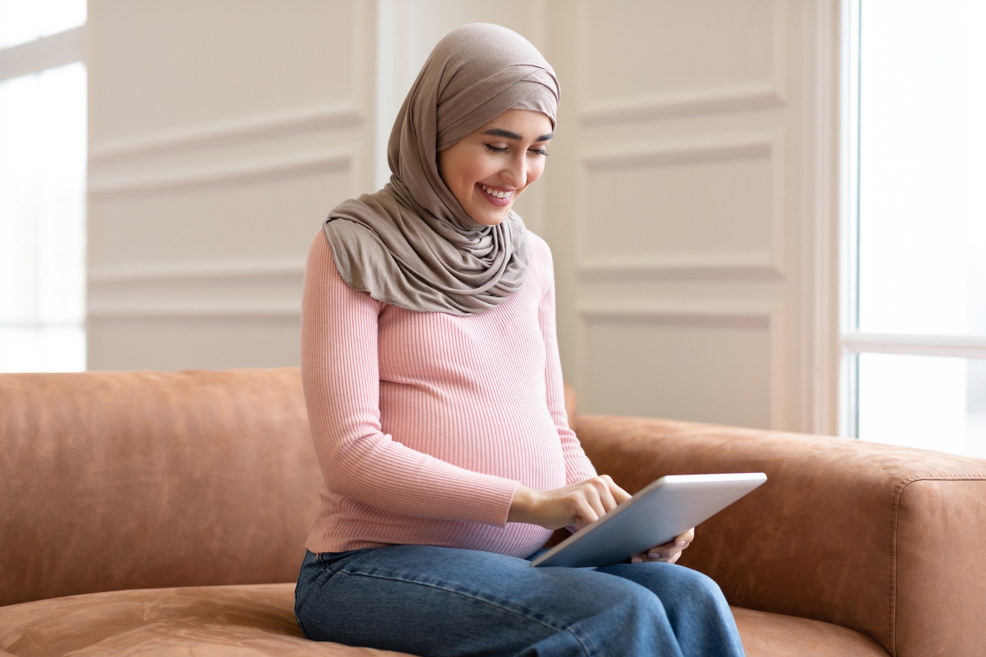 Side View Of Pregnant Muslim Female Using Digital Tablet Computer Browsing Internet Sitting On Sofa At Home. Pregnancy Lifestyle And Modern Gadgets, Technology Concept